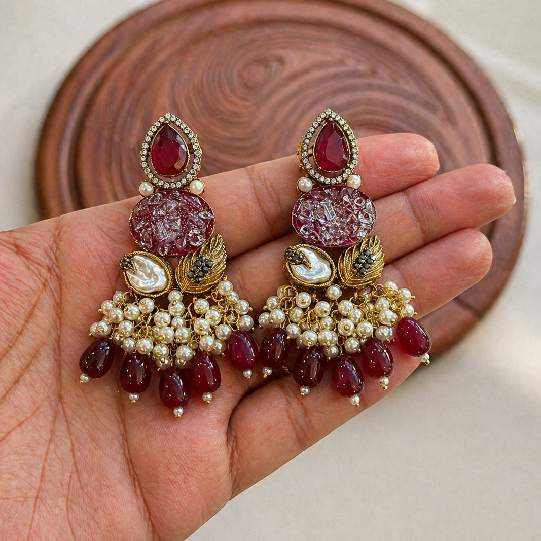 Ornate earrings with red stones and gold accents held in a hand against a neutral background